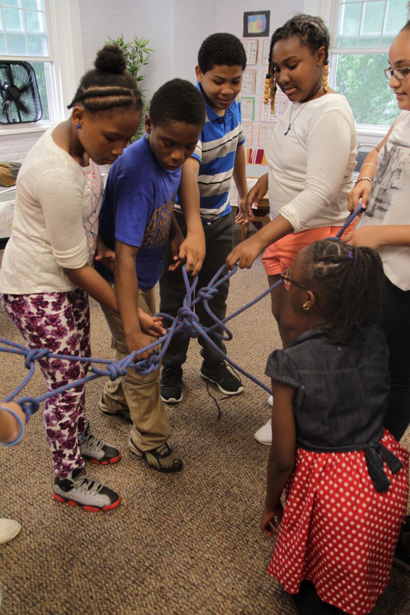 In the image, a group of children is gathered, seemingly engaged in a collaborative activity involving ropes. They are focused on the task at hand, with some holding and manipulating the ropes. The setting appears to be indoors, possibly a classroom or activity room. The children's expressions suggest concentration and teamwork.